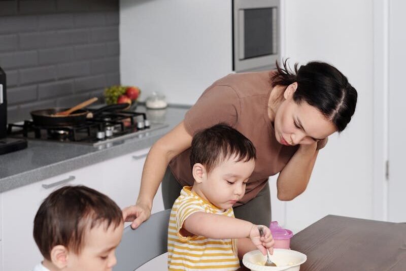 A mother assists her twin boys as they eat at the kitchen table, showcasing family bonding.