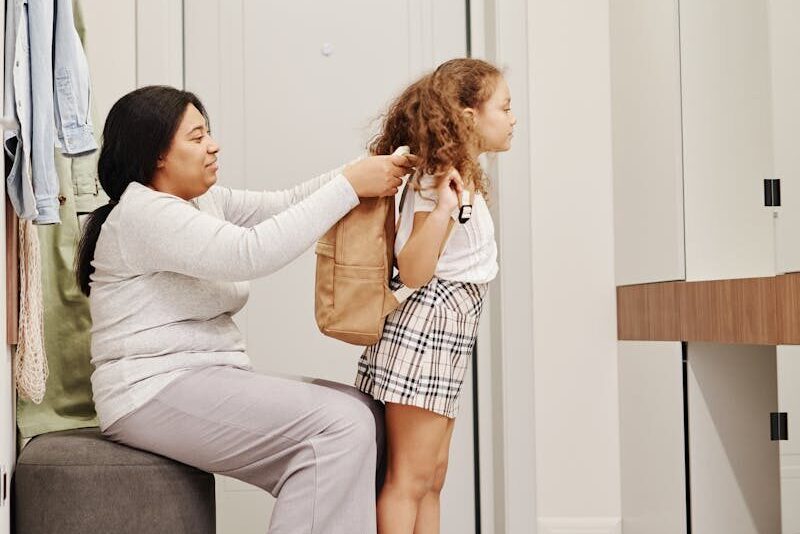A mother helps her daughter put on a backpack indoors, preparing for school.