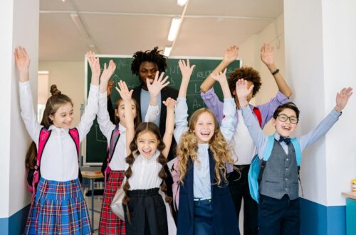 Joyful group of diverse school children raising their hands in an indoor classroom setting.